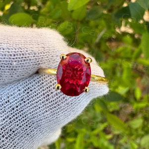 May include: A gold ring with a large, oval-cut, deep red gemstone. The gemstone is held in place by four prongs. The ring's band is plain, polished gold. The background is a soft focus of green foliage.