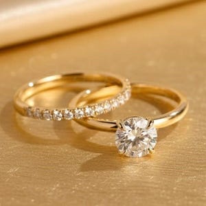 Peut inclure: Deux bagues en or. L'une présente une grosse pierre précieuse ronde et transparente. L'autre est une alliance en or ornée de petites pierres précieuses transparentes. Les bagues sont posées sur une surface dorée.