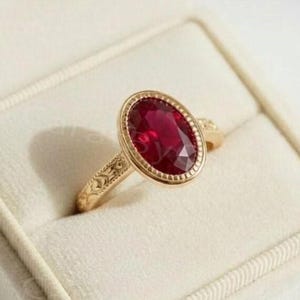 Könnte beinhalten: Ein goldener Ring mit einem ovalen, tiefroten Edelstein. Der Ring ist in einer goldenen Lünette mit einem detaillierten Band gefasst. Er wird in einer weißen Schmuckschatulle mit weichem, samtigem Innenraum präsentiert.