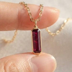 May include: A gold necklace featuring a vertical, rectangular-cut deep red gemstone pendant. The pendant is suspended from a delicate gold chain, showcasing a polished metal setting that frames the vibrant, faceted crimson stone clearly.