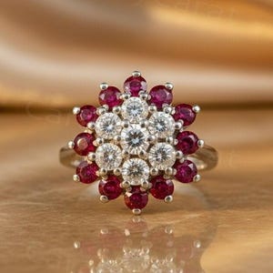 Op de afbeelding: Een zilveren ring met een bloemmotief. De ring heeft een centrale cluster van witte edelstenen, omringd door rode edelstenen. De ring is geplaatst tegen een gouden achtergrond.