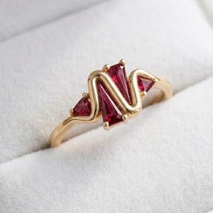 Puede incluir: Un anillo de oro con tres piedras preciosas de color rojo intenso y corte trapezoidal dispuestas en un grupo central. La banda de metal tiene un acabado liso y pulido, y las piedras están engastadas en una montura segura de garras.