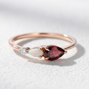 May include: Rose gold ring featuring three gemstones: a small, round diamond, an oval opal, and a pear-shaped garnet. The ring's band is thin and delicate, with the gemstones set close together.