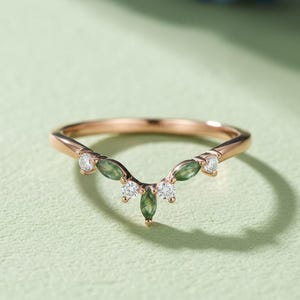 May include: Rose gold ring with a curved design, featuring small green and white gemstones. The ring has a delicate, nature-inspired aesthetic, with the gemstones arranged in a leaf-like pattern. The ring is set against a light green background.