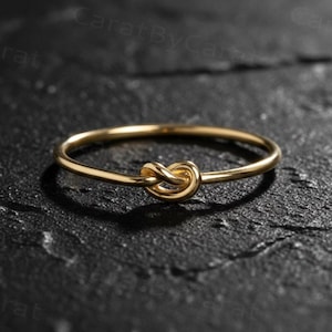 Könnte beinhalten: Ein zarter, goldfarbener Ring mit einem Knotendesign. Der Ring befindet sich auf einer dunklen, strukturierten Oberfläche, die seine schlichte Eleganz hervorhebt. Das Band des Rings ist dünn, und das Knotendetail verleiht diesem Schmuckstück eine einzigartige Note.
