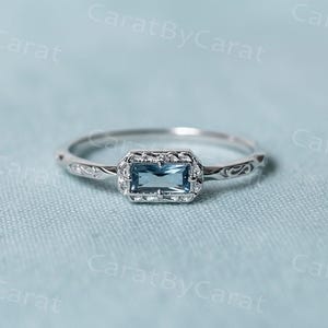 Peut inclure: Bague en argent avec une pierre précieuse rectangulaire bleu clair, sertie dans un cadre rectangulaire. La monture présente un motif floral gravé. La bague est posée sur un fond bleu clair.