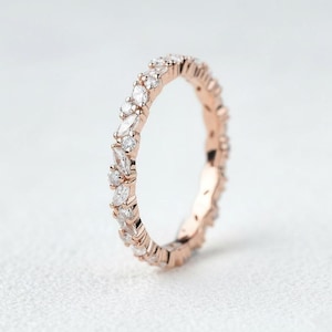 May include: Rose gold ring featuring a continuous band of alternating round and marquise-cut clear gemstones. The ring is set against a soft, white background. A delicate piece of jewellery.