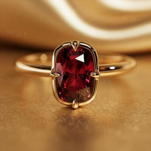 Puede incluir: Un anillo de oro con una gran piedra preciosa roja oscura rectangular. La piedra preciosa está engastada en un bisel de oro y tiene una superficie facetada. El anillo está sobre una superficie dorada.