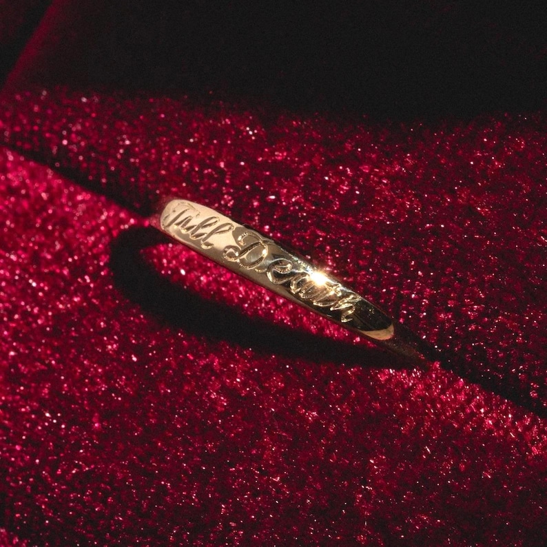 May include: A gold ring with the inscription "Till Death" engraved on the band. The ring is set against a deep red, textured background, creating a contrast that highlights the ring's design.