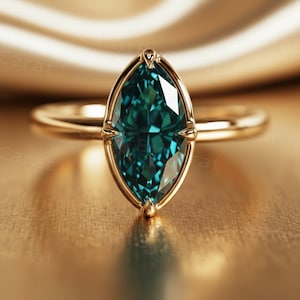 May include: A gold ring featuring a marquise-cut, teal gemstone. The gemstone is set in a gold bezel, with a polished gold band. The ring is presented on a reflective, golden surface.