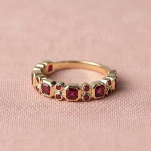 Pode incluir: Um anel de ouro com uma série de pedras preciosas vermelhas quadradas e redondas. O anel é apresentado sobre um fundo rosa suave, destacando a cor vibrante das pedras e o design intrincado do anel. Uma joia.