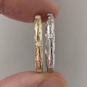 May include: Two rings, one gold and one silver, are held between fingers. Both rings feature a repeating pattern of stars and small gemstones. The gold ring has a warm, yellow hue, while the silver ring has a bright, reflective finish.
