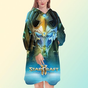 May include: A hooded blanket featuring a detailed graphic from the video game StarCraft II: Legacy of the Void. The design showcases a futuristic helmet in shades of blue, gold, and green, with the game's logo at the bottom. The blanket is a wearable, oversized design.