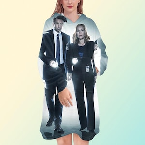 May include: A hooded blanket featuring a printed image of two people in suits holding flashlights. The image is a promotional graphic for a television show. The blanket is white with a light blue and gray background.