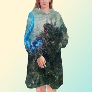 Può includere: Una coperta indossabile con cappuccio oversize con un design Godzilla. Il design mostra Godzilla in un ambiente urbano con effetti di energia blu. La coperta è di colore scuro con cappuccio e maniche lunghe.