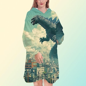 May include: A hooded blanket with a Godzilla design. The design depicts Godzilla above a city, set against a cloudy sky. The blanket is mainly teal and blue, with a white interior.