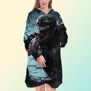 May include: Black hooded blanket featuring a detailed graphic of the Venom character. The design showcases Venom's face with sharp teeth and a long red tongue. The blanket has a soft, plush appearance and includes pockets.