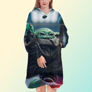 May include: A hooded sweatshirt featuring a graphic of a green alien character with large eyes and ears. The design includes a dark cloak and a background with blue and green tones. The sweatshirt has a v-neck and a relaxed fit.