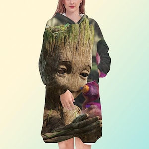 May include: A hooded sweatshirt featuring a detailed graphic of Groot from Guardians of the Galaxy. The design showcases Groot's wooden texture and facial features, with a purple creature in his arms. The sweatshirt has a white interior and a long, oversized fit.