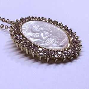 May include: Gold-toned necklace featuring an oval pendant with a carved white cameo design. The pendant is surrounded by a halo of small, clear gemstones. The necklace chain is delicate and gold-colored, creating an elegant accessory.