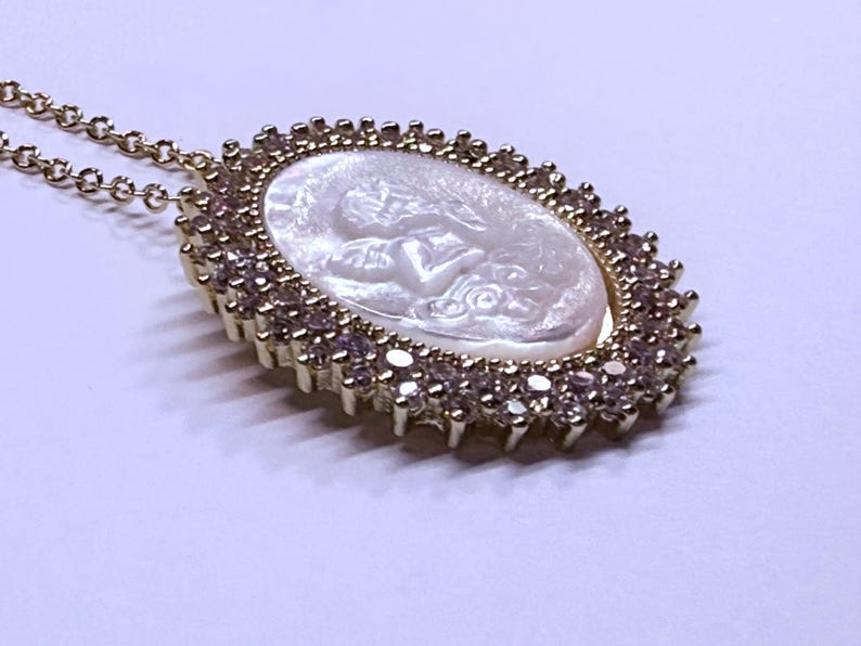 May include: Gold-toned necklace featuring an oval pendant with a carved white cameo design. The pendant is surrounded by a halo of small, clear gemstones. The necklace chain is delicate and gold-colored, creating an elegant accessory.