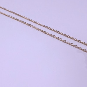 May include: Close-up of two delicate gold-colored chain necklaces. The chains are composed of small, evenly spaced oval links. The background is a soft, light purple color, providing a subtle contrast to the gold chains.
