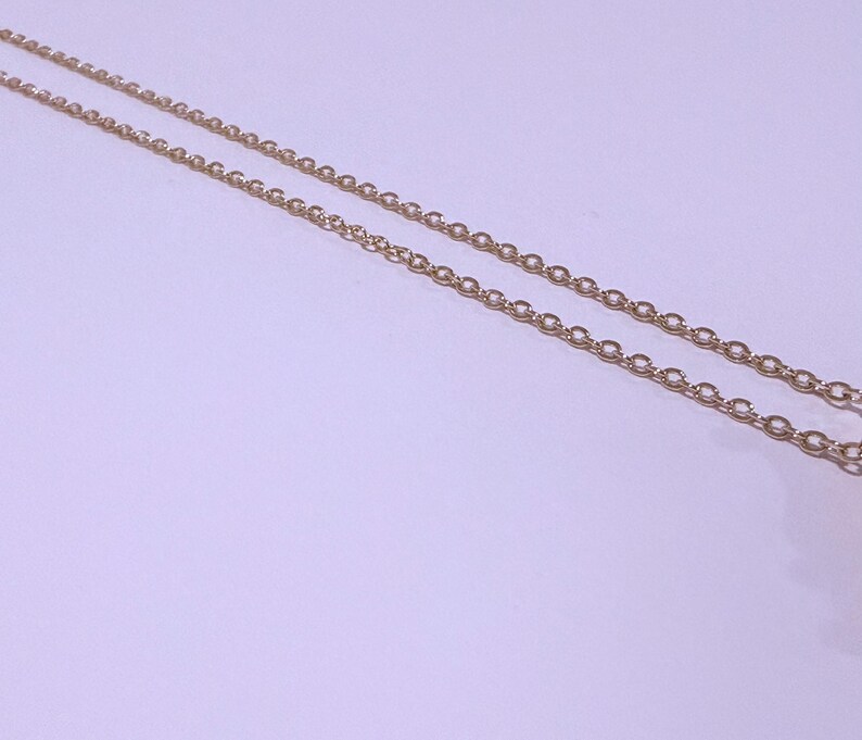 May include: Close-up of two delicate gold-colored chain necklaces. The chains are composed of small, evenly spaced oval links. The background is a soft, light purple color, providing a subtle contrast to the gold chains.