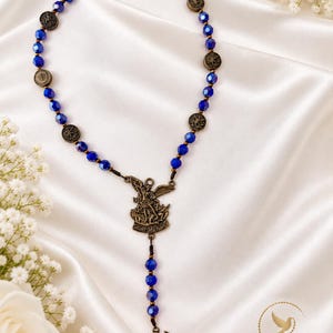 May include: A rosary featuring deep blue faceted beads interspersed with small, round, bronze-toned metal medallions. A central bronze-toned centerpiece depicts Saint Michael, and a winged sword pendant hangs at the bottom of the strand.
