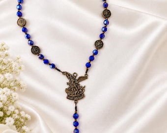 Peut inclure: Un chapelet composé de perles facettées bleu profond alternant avec de petits médaillons métalliques de couleur bronze. Une pièce centrale en bronze représente Saint Michel, et un pendentif en forme d'épée ailée termine le chapelet.