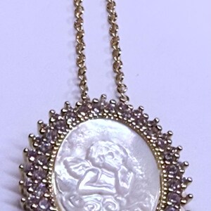 May include: Gold-toned necklace featuring a pendant with a carved white cameo of an angel. The pendant is surrounded by a halo of small, sparkling stones. The necklace chain is delicate and also gold-toned.