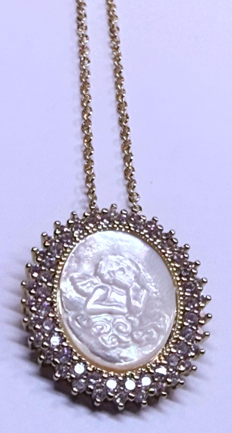 May include: Gold-toned necklace featuring a pendant with a carved white cameo of an angel. The pendant is surrounded by a halo of small, sparkling stones. The necklace chain is delicate and also gold-toned.