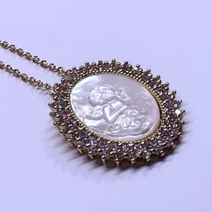May include: Gold-toned necklace featuring an oval pendant with a carved cherub design on a mother-of-pearl background. The pendant is framed by a halo of small, clear and pink gemstones, and is suspended from a delicate chain.