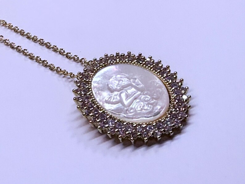 May include: Gold-toned necklace featuring an oval pendant with a carved cherub design on a mother-of-pearl background. The pendant is framed by a halo of small, clear and pink gemstones, and is suspended from a delicate chain.