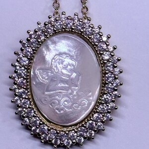 May include: Gold-toned necklace with an oval pendant. The pendant features a carved angel design within a mother-of-pearl oval, surrounded by a halo of clear gemstones. The chain is delicate and gold-colored, suitable for everyday wear.