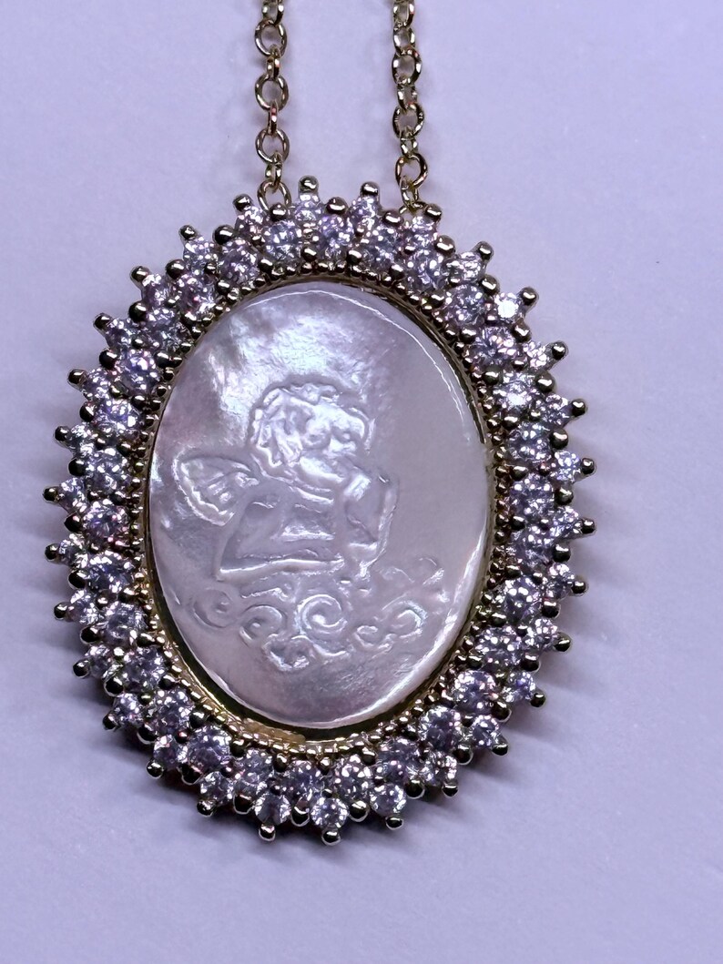 May include: Gold-toned necklace with an oval pendant. The pendant features a carved angel design within a mother-of-pearl oval, surrounded by a halo of clear gemstones. The chain is delicate and gold-colored, suitable for everyday wear.