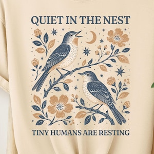 Peut inclure: Un t-shirt beige à manches courtes avec une impression graphique de deux oiseaux perchés sur des branches avec des fleurs, des étoiles et un croissant de lune. Le texte QUIET IN THE NEST et TINY HUMANS ARE RESTING est écrit en bleu foncé.