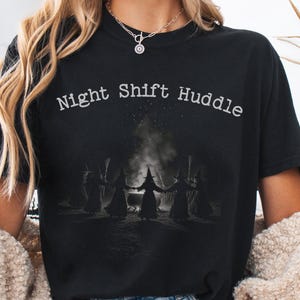 May include: A black short-sleeve t-shirt featuring the white text Night Shift Huddle above a graphic of five figures in pointed hats standing in a circle around a steaming cauldron. The shirt has a crew neckline and a relaxed fit design.
