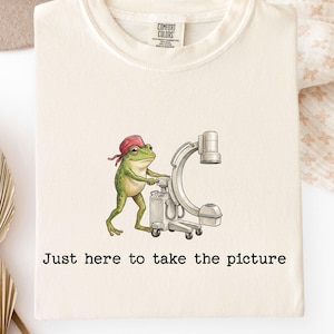 May include: A cream-colored t-shirt featuring a graphic of a green frog wearing a red head covering and operating a medical C-arm imaging machine. Below the illustration, the text reads, Just here to take the picture, in black lettering.
