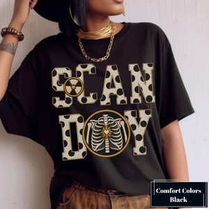 May include: A black t-shirt featuring the text SCAN DOY in large, beige, polka-dotted varsity-style letters. The letter A contains a circular radiation symbol, and the letter O is replaced by a circular graphic of a human rib cage with a heart.
