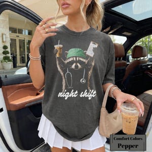 Puede incluir: Una camiseta gris carbón con un gráfico de un mapache que lleva un gorro quirúrgico verde y un estetoscopio. El mapache sostiene una taza de café y un portapapeles. El texto "night shift" está impreso en blanco debajo del gráfico. La camiseta está etiquetada como "Comfort Colors" y "Pepper".
