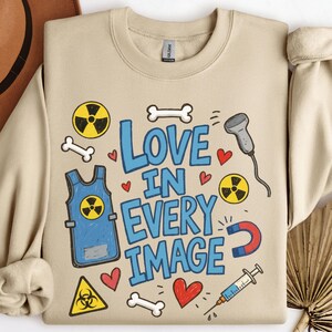 Może przedstawiać: Beżowa bluza z okrągłym dekoltem z niebieskim napisem "LOVE IN EVERY IMAGE" otoczonym ilustracjami fartucha ołowianego, symboli promieniowania, kości, czerwonego serca, głowicy USG, magnesu podkowiastego i strzykawki.