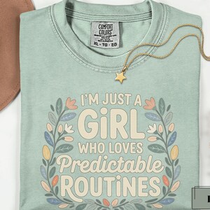 May include: A light green t-shirt with the text "I'M JUST A GIRL WHO LOVES PREDICTABLE ROUTINES" in a floral design. A gold star pendant necklace is draped across the shirt. The shirt is a size XL-TG-EG.