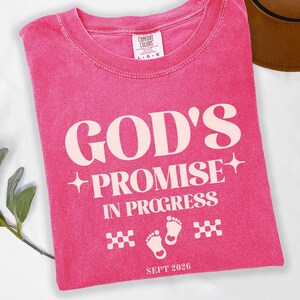 Puede incluir: Una camiseta rosa con las palabras "GOD'S PROMISE IN PROGRESS" en color crema. La camiseta también presenta huellas de bebé y la fecha "SEPT 2026". La camiseta está doblada y tiene un aspecto vintage.