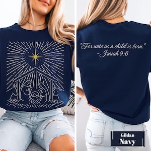 Puede incluir: Una sudadera azul marino con un gráfico de líneas blancas de una escena de natividad y una estrella amarilla brillante en el frente. La parte posterior muestra el texto blanco: "For unto us a child is born." - Isaiah 9:6. Una etiqueta dice Gildan Navy.