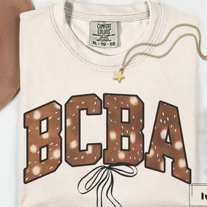 Könnte beinhalten: Elfenbeinfarbenes T-Shirt mit den Buchstaben "BCBA" in einem braunen, gefleckten Muster, umrandet von dunklerem Braun. Eine goldene Sternanhänger-Halskette und ein brauner Hut sind sichtbar. Das Shirt hat ein "Comfort Colors"-Etikett.