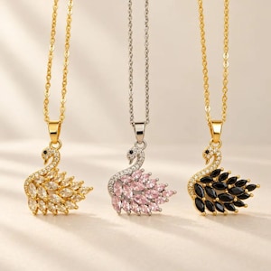 May include: Three necklaces featuring swan-shaped pendants suspended from delicate chains. The left pendant has clear crystals, the centre has pink crystals, and the right has black crystals. Each swan is accented with small clear stones along the neck.