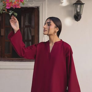 Puede incluir: Túnica burdeos de manga larga y cuello en V. La túnica tiene mangas acampanadas y un corte holgado. La modelo lleva la túnica al aire libre, cerca de una ventana y plantas con flores. La tela parece suave.