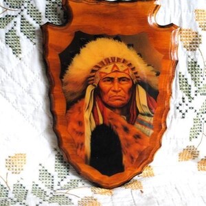 May include: A framed painting of a Native American man wearing a headdress with feathers. The painting is on a wooden background with a carved edge.