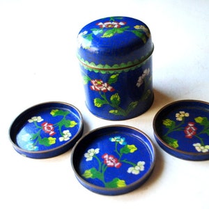 May include: A blue enamel container and three matching coasters. The set features a floral design with red, white, and green accents. The container is cylindrical with a lid, and the coasters are round with a bronze trim.