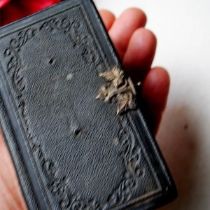 May include: A small, black leather-bound book with an ornate design and a metal clasp. The book is closed and appears to be very old.